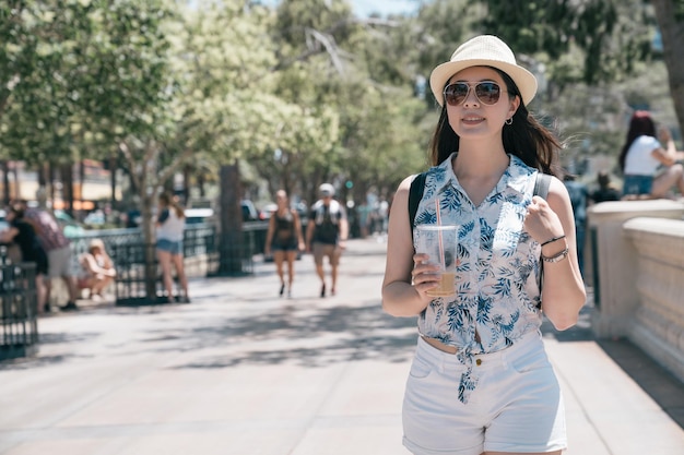 Lächelnder Hipster-Reisender in stilvoller Kleidung, der am sonnigen Wochenendtag den Morgenkaffee zum Mitnehmen genießt, während er auf der Straße der Stadt spazieren geht. Fröhliche Touristinnen mit Rucksack entspannen sich auf dem Boulevard von Las Vegas.