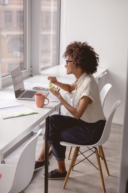 Lächelnder hübscher Arbeiter, der am Arbeitsplatz in der Nähe des Fensters sitzt