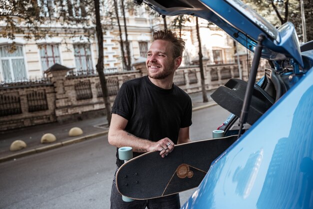 Lächelnder hübscher blonder männlicher Skater, der Longboard in den Kofferraum des Autos legt und jemanden auf dem Stadthintergrund sieht. Extremsportarten. Urbane Hobbys. Freizeitkonzept. Freizeit, Outdoor-Konzept.