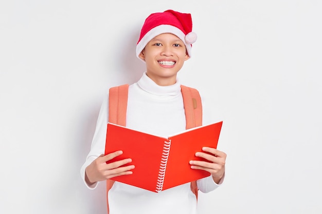 Lächelnder hübscher junger asiatischer Student in der Weihnachtsmütze und in der zufälligen Kleidung mit dem Rucksack, der das Buch lokalisiert auf weißem Hintergrund hält