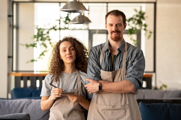 Lächelnder hübscher junger bärtiger Mann im Jeanshemd, der auf modernem Laptop beim Arbeiten im Café tippt