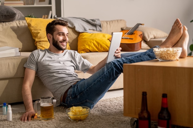 Lächelnder hübscher junger Mann in der Freizeitkleidung, die mit Füßen auf Kaffeetisch sitzt und mit Freunden online zu Hause Isolation plaudert