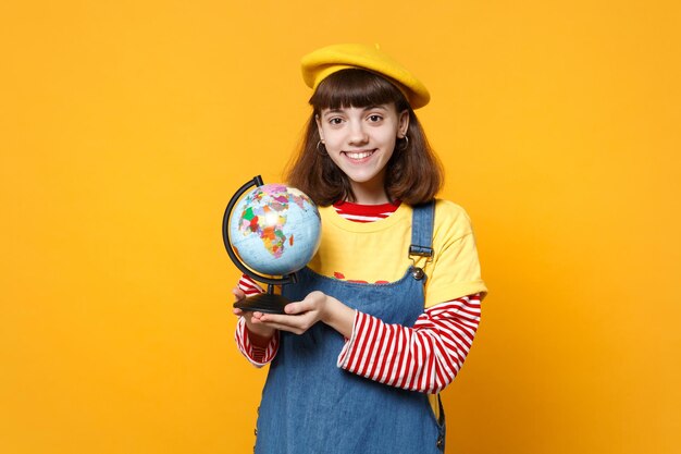 Lächelnder hübscher Teenager in französischer Baskenmütze und Denim-Sommerkleid, der die Erdkugel isoliert auf gelbem Wandhintergrund im Studio hält. Menschen aufrichtige Emotionen, Lifestyle-Konzept. Mock-up-Kopienbereich.