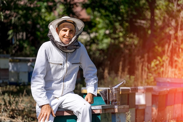 Lächelnder Imker in weißer Uniform Mann sitzt in der Nähe von Arbeitsgeräten und Bienenstöcken