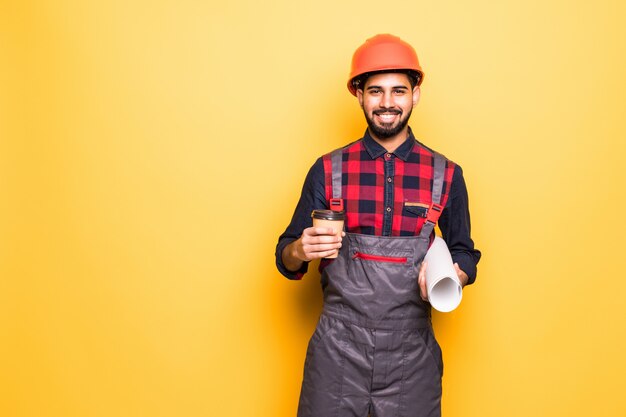 Lächelnder indischer Ingenieur in der Sicherheitsweste, die Blaupausen und Kaffee hält, um auf gelbem Raum isoliert zu gehen