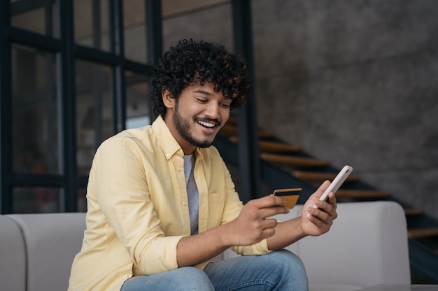 Lächelnder indischer Mann, der eine Kreditkarte mit Handy beim Online-Scheckkartenguthaben hält