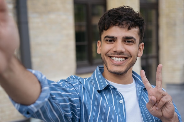 Lächelnder indischer Mann mit Handy, der ein Selfie mit Victory-Zeichen auf der Straße macht