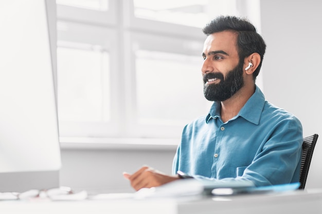 Lächelnder indischer Mann mit Ohrhörer arbeitet an einem hellen Bürodesk