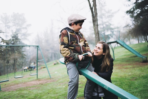 Lächelnder Junge, der mit seiner Mutter auf einem Spielplatz in einer Wippe spielt. Freizeitgenuss im Park.