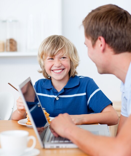 Lächelnder Junge, der während sein Vater mit einem Laptop frühstückt