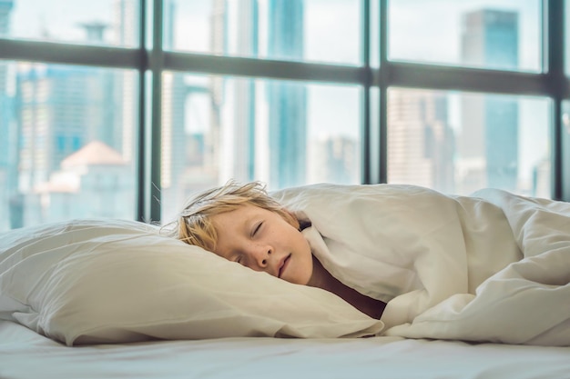 Lächelnder Junge im Bett, der in einer Großstadt im Hintergrund von Wolkenkratzern aufwacht. Kindergesundheit im Großstadtkonzept