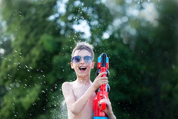 Lächelnder Junge in den Gläsern spielt mit Wasserpistole am heißen sonnigen Sommertag