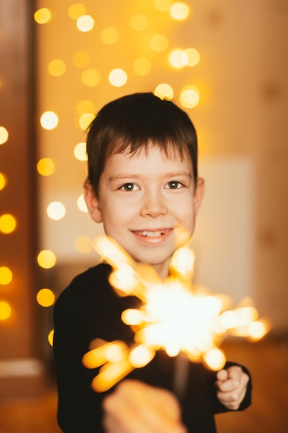 Lächelnder Junge mit Feuerwerk in seinen Händen vor dem Hintergrund einer gelben Weihnachtsgirlande Glückliches Kind mit Wunderkerzen auf Silvesterfeier