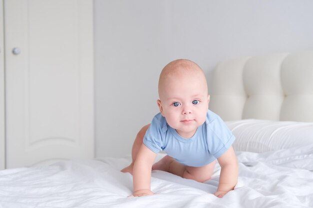 lächelnder Junge mit großen blauen Augen in einem Bodysuit lernt, auf weißer Bettwäsche zu kriechen Gesundes neugeborenes Baby