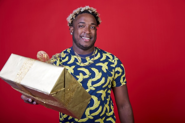 Lächelnder junger Afroamerikaner gibt ein Geschenk in einem goldenen Paket auf rotem Hintergrund