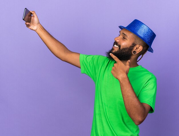 Lächelnder junger Afroamerikaner mit Partyhut macht Selfie-Punkte am Telefon