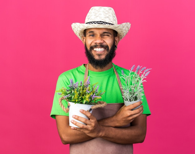 Lächelnder junger afroamerikanischer Gärtner mit Gartenhut, der Blumen im Blumentopf hält und überquert, isoliert auf rosa Wand