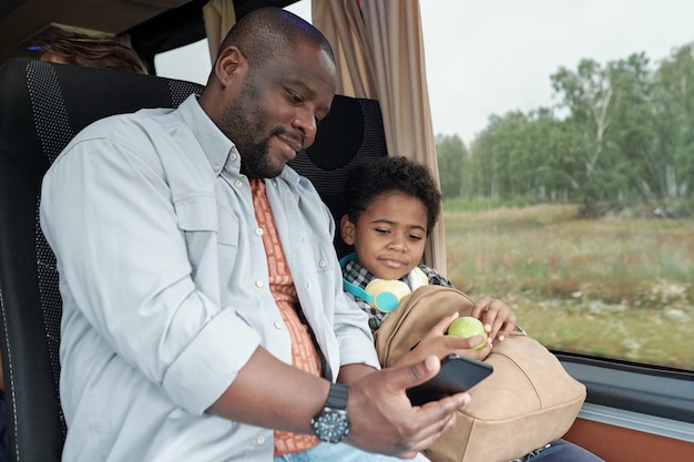 Lächelnder junger afroamerikanischer Vater mit Smartphone beim Ansehen von Videos mit Sohn während der Busfahrt