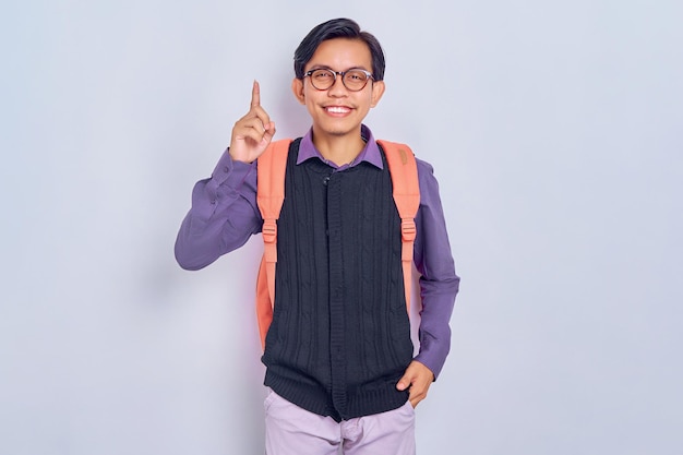 Lächelnder junger asiatischer männlicher Student vom College in lässiger Kleidung mit Rucksack, der mit dem Finger nach oben zeigt und eine Idee denkt, die auf grauem Hintergrund isoliert ist Bildung in High School University College-Konzept