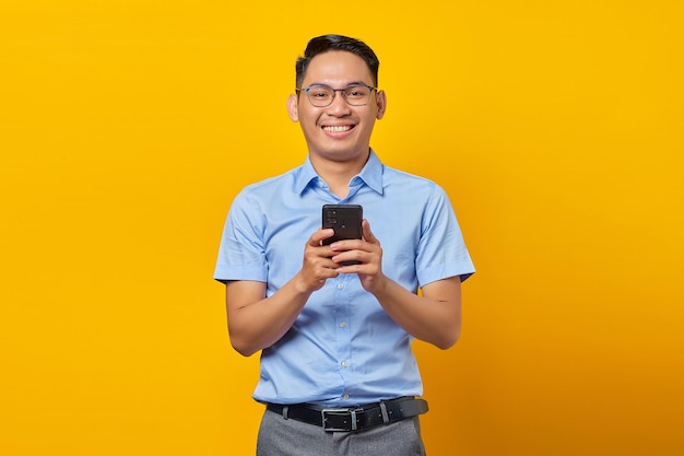Lächelnder junger asiatischer Mann Asiate mit Brille antwortet auf eingehende Nachrichten auf dem Handy isoliert auf gelbem Hintergrund Geschäftsmann- und Unternehmerkonzept