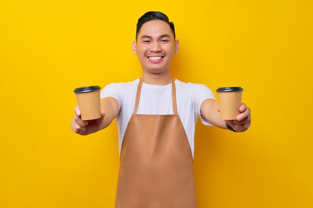 Lächelnder junger asiatischer Mann, Barista, Barman-Mitarbeiter, der eine braune Schürze trägt, arbeitet im Café und gibt eine Tasse Kaffee oder Tee zum Mitnehmen, die auf gelbem Hintergrund isoliert ist