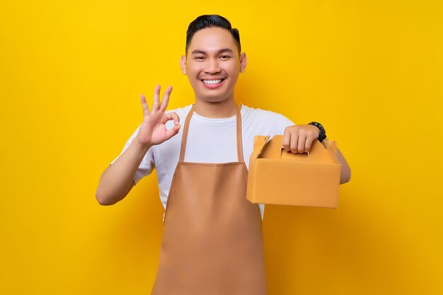 Lächelnder junger asiatischer Mann, Barista, Barman-Mitarbeiter, der eine braune Schürze trägt, arbeitet in einem Café, das eine Papiertüte mit Kuchen zum Mitnehmen hält und eine okay-Geste auf gelbem Hintergrund zeigt. Startup-Konzept für kleine Unternehmen