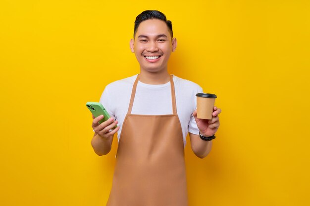 Lächelnder junger asiatischer Mann Barista Barman-Mitarbeiter mit brauner Schürze arbeitet im Café mit mobilem Phoe und Pappbecher Kaffee oder Tee isoliert auf gelbem Hintergrund Kleinunternehmensgründung