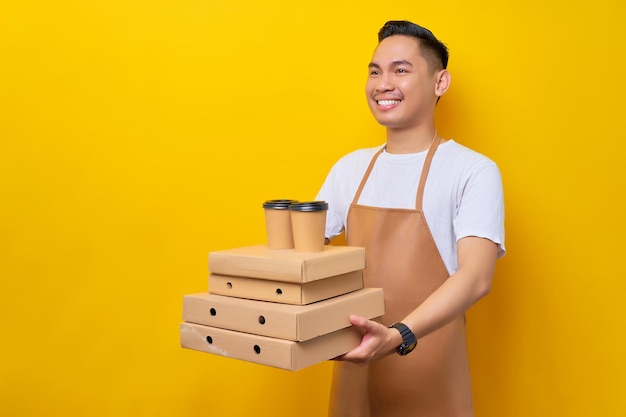 Lächelnder junger asiatischer Mann Barista Barman-Mitarbeiter mit brauner Schürze, der im Café arbeitet und Liefertassen-Pizza-Karton-Flatbox auf gelbem Hintergrund gibt Kleines Unternehmensgründungskonzept