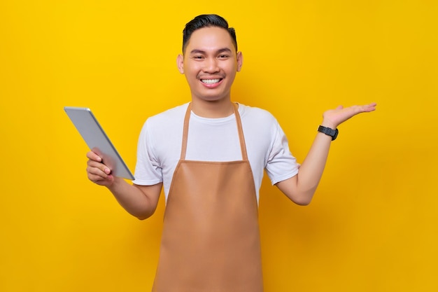 Lächelnder junger asiatischer Mann Barista Barman-Mitarbeiter mit brauner Schürze, der im Café mit digitalem Tablet arbeitet und mit der Hand auf gelbem Hintergrund im Kopierbereich zeigt Kleinunternehmensgründung