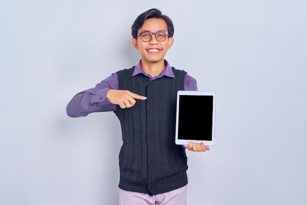 Lächelnder junger asiatischer Mann in lässigem Hemd und Weste, der mit dem Finger auf den leeren Bildschirm des Tablet-PC-Computers zeigt, der auf weißem Hintergrund isoliert ist People-Lifestyle-Konzept
