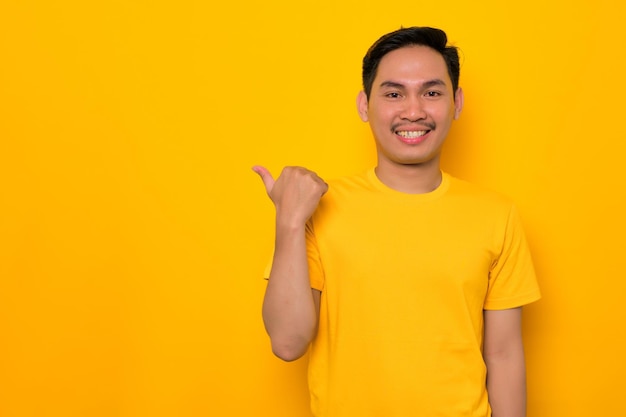 Lächelnder junger asiatischer Mann in lässigem T-Shirt, der mit dem Daumen zur Seite auf den Kopierbereich zeigt, der auf gelbem Hintergrund isoliert ist Tolles Angebotskonzept