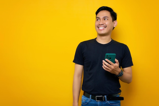Lächelnder junger asiatischer Mann in legerem T-Shirt, der ein Handy hält und beiseite schaut, isoliert auf gelbem Hintergrund Lifestyle-Konzept für Menschen