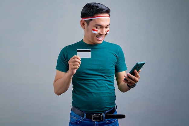 Lächelnder junger asiatischer Mann in legerem T-Shirt mit Handy und Kreditkarte isoliert auf grauem Hintergrund indonesisches Unabhängigkeitstag-Feierkonzept