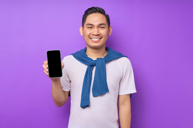 Lächelnder junger asiatischer Mann in legerer Kleidung mit Handy mit leerem Bildschirm isoliert auf violettem Hintergrund Werbung für neues Mockup-Konzept für mobile Apps