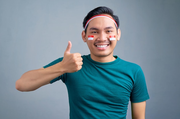 Lächelnder junger asiatischer mann mit daumen nach oben geste isoliert auf grauem hintergrund indonesisches unabhängigkeitstag-feierkonzept