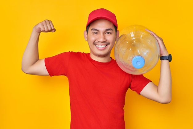 Lächelnder junger asiatischer Mann mit rotem T-Shirt und einheitlichem Mitarbeiter arbeitet als Händlerkurier, der eine Wasserflasche mit isoliertem Muskel auf gelbem Hintergrund hält Professionelles Lieferservice-Konzept