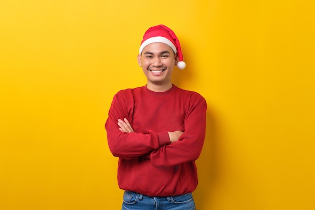 Lächelnder junger asiatischer Mann mit Weihnachtsmütze, der mit verschränkten Armen in die Kamera schaut, über gelbem Studiohintergrund Feier Weihnachtsfeiertag und Neujahrskonzept