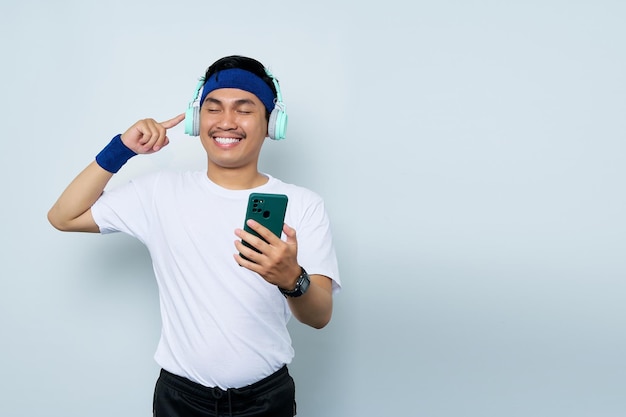 Lächelnder junger asiatischer Mann, sportlicher Fitness-Trainer mit blauem Stirnband und weißem T-Shirt. Hören Sie Musik mit Kopfhörern und verwenden Sie das Handy, das über weißem Hintergrund isoliert ist