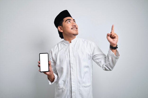 Foto lächelnder junger asiatischer muslimischer mann in weißer kleidung, der ein smartphone hält, das einen bildschirm zeigt, der aufrecht steht