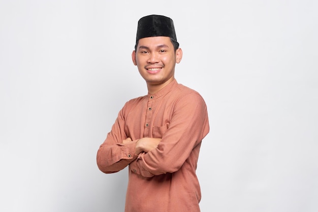 Lächelnder junger asiatischer muslimischer Mann verschränkte die Arme und sah selbstbewusst isoliert auf weißem Hintergrund aus