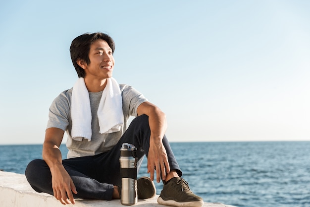 Lächelnder junger asiatischer Sportler, der am Meer stillsteht