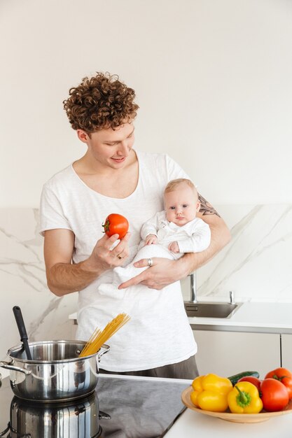 Lächelnder junger attraktiver Vater, der Nudeln kocht, während er seinen kleinen Sohn in der Küche zu Hause hält