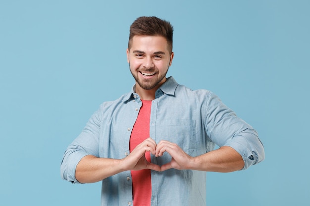 Lächelnder junger bärtiger Kerl der 20er Jahre im Freizeithemd posiert isoliert auf pastellblauem Hintergrund Studioporträt. People-Lifestyle-Konzept. Mock-up-Kopienbereich. Formherz mit Händen, Herzformzeichen zeigen.