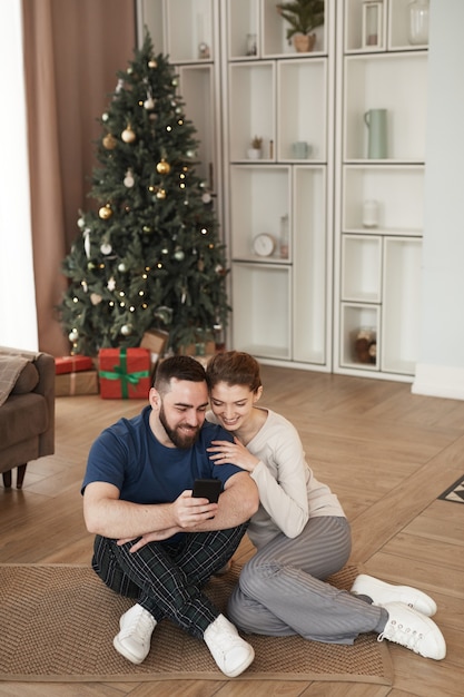 Lächelnder junger bärtiger Mann sitzt auf dem Boden im Wohnzimmer mit Weihnachtsbaum und zeigt Video auf ...