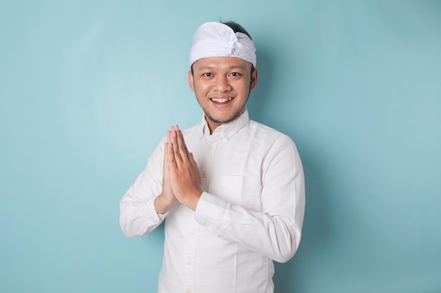 Lächelnder junger balinesischer Mann mit Udeng oder traditionellem Stirnband und weißem Hemd, der Gruß oder Namaste einzeln auf blauem Hintergrund gestikuliert