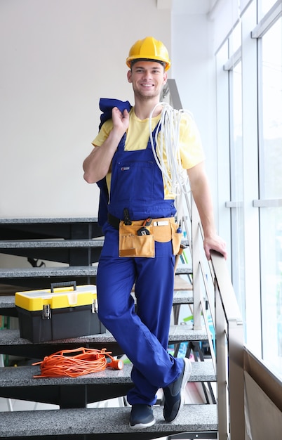 Lächelnder junger Elektriker mit Kabelbündel und Werkzeugkasten, der drinnen auf Treppen steht