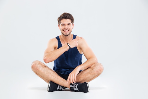 Lächelnder junger Fitness-Mann, der mit gekreuzten Beinen sitzt und auf weißem Hintergrund zeigt