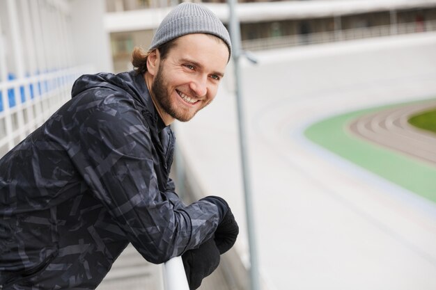 Lächelnder junger fitter Sportler, der sich nach dem Training im Stadion ausruht und sich auf eine Schiene stützt