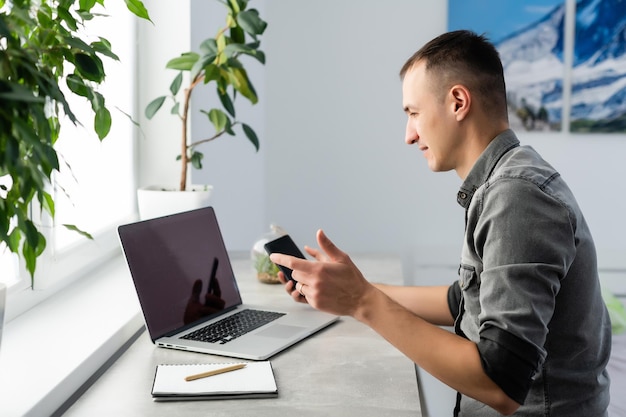 Lächelnder junger Freiberufler mit Laptop, Online-Studium, Arbeit von zu Hause aus, glücklicher Gelegenheitstyp, der auf einem PC-Notebook tippt, im Internet surft, entfernten Job genießt, am Tisch sitzt.