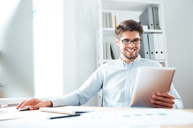 Lächelnder junger Geschäftsmann, der am Tisch sitzt und Tablet-Computer im Büro benutzt